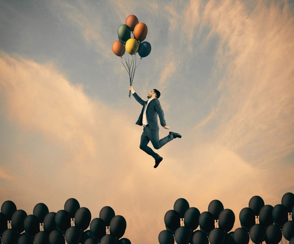 Homme en costume s'envolant avec des ballons de baudruche colorés au-dessus de ballons noirs, symbolisant la liberté et le changement de vie